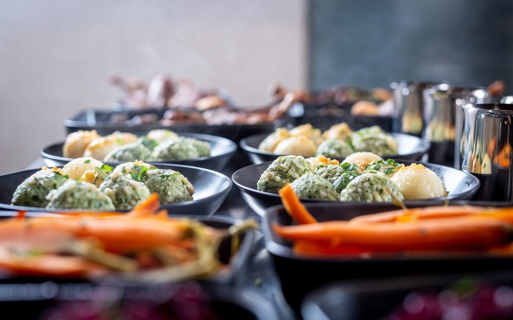 EVENTGIESSEREI Catering: Das Bild zeigt mehrere dunkle Schalen, gefüllt mit dampfenden Möhren, Braten und weiteren Lebensmitteln.