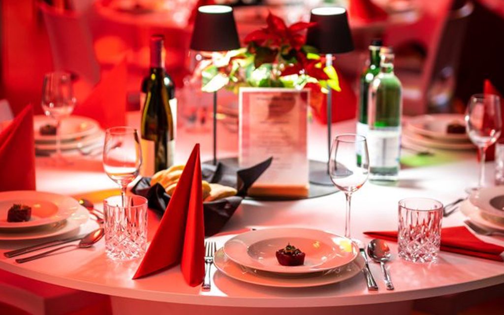 Ein festlich gedeckter Tisch für eine Weihnachtsfeier in Leipzig in der EVENTGIESSEREI. Auf dem weißen runden Tisch stehen weißes Porzellangeschirr, Weingläser, verschiedene Weinflaschen, Dekoration und LED-Tischlampen.