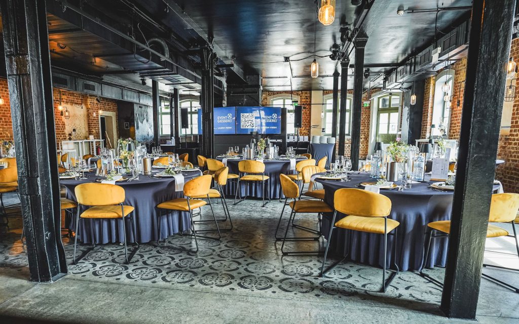 Innenraum der Eventlocation EVENTGIESSEREI Leipzig mit goldenen Samtstühlen, dunklen Tischen und Bodenmosaiken.