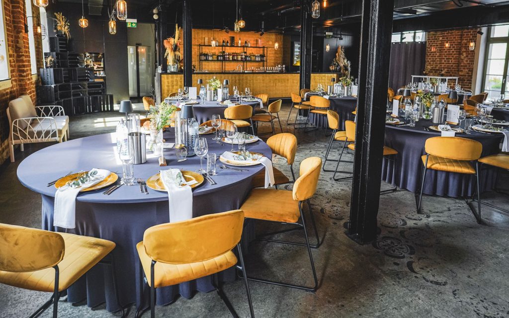 Innenraum der Location EVENTGIESSEREI mit runden Banketttischen, dunkel behusst, und goldenen Samtstühlen und im Hintergrund der goldenen Bar.