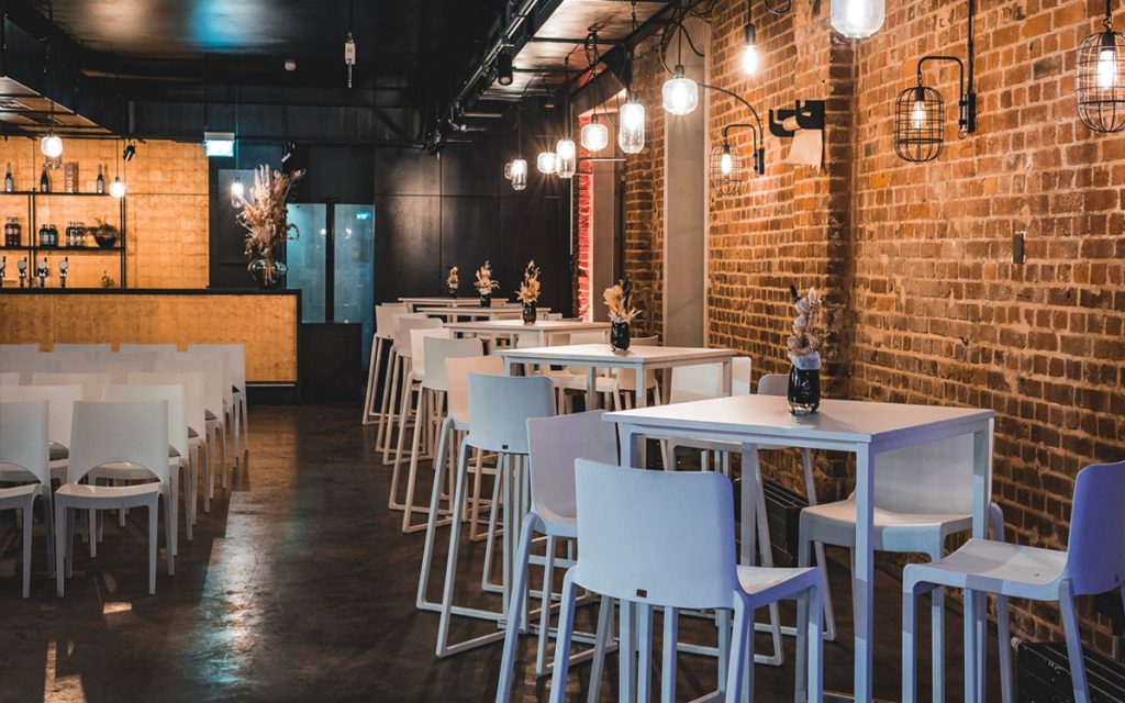 Setup für Marken-Events & Produktlaunches: weiße Stehtische und Barhocker, im Hintergrund rote Ziegelwand mit Lampen im Industrial Style