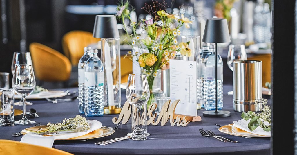 Für eine Hochzeit eingedeckter Tisch mit dunkler Husse, goldenen Platztellern, feiner Tischdeko und Getränken